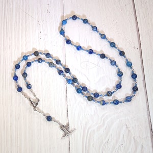May include: A blue lapis lazuli beaded necklace with a silver pendant in the shape of a bird. The necklace is on a white wooden surface.