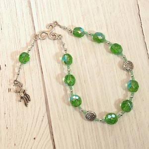 Puede incluir: Una pulsera de cuentas verdes con detalles plateados. La pulsera presenta grandes cuentas verdes facetadas y cuentas plateadas más pequeñas. Un dije plateado en forma de ciervo está adjunto. La pulsera se exhibe sobre una superficie de madera clara.