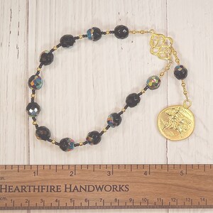 Pode incluir: Um mala de contas pretas com detalhes iridescentes e um amuleto dourado. O mala tem 38 cm de comprimento e uma borla dourada.
