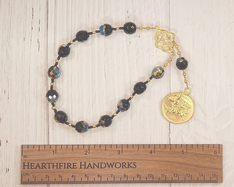 Pode incluir: Um mala de contas pretas com detalhes iridescentes e um amuleto dourado. O mala tem 38 cm de comprimento e uma borla dourada.