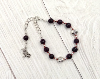 May include: A silver chain bracelet with dark purple beads and silver charms. The charms are Celtic knot designs. A silver crow charm hangs from the chain.