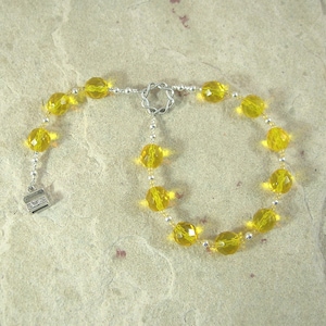 May include: A bracelet featuring faceted, yellow beads strung on a silver chain. The bracelet has a decorative clasp and a small, square charm. The beads are evenly spaced and reflect light, creating a sparkling effect. The jewelry is displayed on a neutral surface.