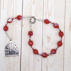 May include: A rosary with faceted red beads and silver-toned accents. The rosary features a decorative silver-toned pendant shaped like a door. The beads are strung on a silver-toned chain.