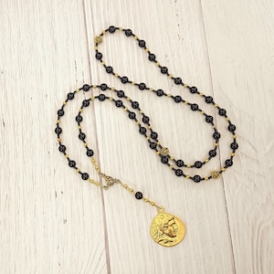 Puede incluir: Un collar de cuentas negras y doradas con un colgante de moneda de oro que presenta un perfil de un rostro.