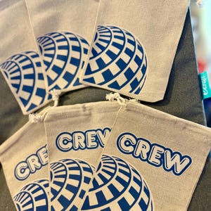 Pode incluir: Várias sacolas cinza claro com cordão, com um design de globo azul e branco e a palavra "CREW" impressa em azul. As sacolas são feitas de tecido natural.