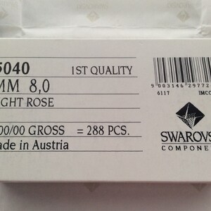 Pode incluir: Uma caixa branca com texto preto que diz "5040 MM 8,0 LIGHT ROSE 2/00/00 GROSS = 288 PCS. Made in Austria" e um c&oacute;digo de barras com o texto "9 003146 297724> 6117 IMCOMO". A caixa tamb&eacute;m tem um logotipo Swarovski com o texto "SWAROVSKI COMPONENTS".