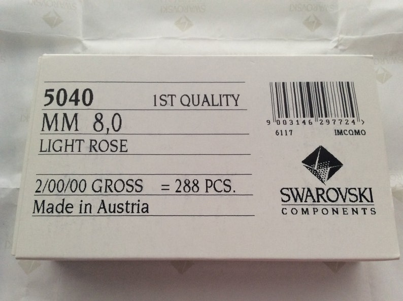 Pode incluir: Uma caixa branca com texto preto que diz "5040 MM 8,0 LIGHT ROSE 2/00/00 GROSS = 288 PCS. Made in Austria" e um c&oacute;digo de barras com o texto "9 003146 297724> 6117 IMCOMO". A caixa tamb&eacute;m tem um logotipo Swarovski com o texto "SWAROVSKI COMPONENTS".