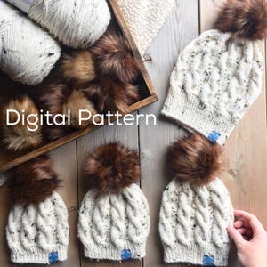 Puede incluir: Tres gorros de punto blanco con pompones de piel sintética marrón. Los gorros tienen un patrón de punto de cable y están colocados sobre una superficie de madera. El texto "Digital Pattern" es visible en la imagen.