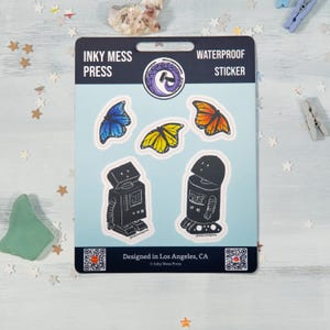 May include: A sheet of waterproof stickers featuring two robots, three butterflies, and the text "Inky Mess Press" and "Waterproof Sticker". The butterflies are blue, orange, and yellow. The robots are black and white.