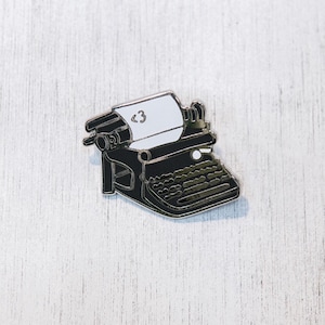 May include: Black and white enamel pin of a vintage typewriter with the text "<3" on the paper.