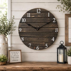 May include: A large round wall clock constructed from horizontal dark brown stained wooden planks. The clock face features white Arabic numerals from 1 to 12 and two thin black metal hands centered on the dial to indicate the time.
