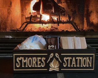 May include: A wooden box with the words "S'mores Station" on the front. The box is divided into three compartments, each containing ingredients for making s'mores: mini marshmallows, chocolate bars, and graham crackers.