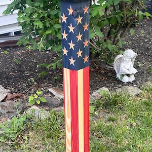 May include: A wooden solar post light painted with the American flag design. The top section is navy blue with gold stars, and the bottom section has red and yellow stripes. The light is square and has a black top.