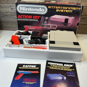 Może przedstawiać: Zestaw Nintendo Entertainment System Action Set w stylu vintage, zawierający konsolę, pistolet świetlny Zapper i broszurę Control Deck. Pudełko zawiera logo Nintendo i zdjęcia produktów. Zestaw jest w oryginalnym opakowaniu.