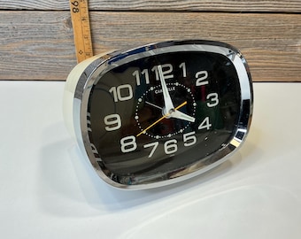May include: A vintage Carvelle alarm clock with a black face and white numerals. The clock has a chrome bezel and a cream-colored body. The clock face features white hands and a yellow second hand.