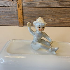 May include: A white porcelain butter dish with a small porcelain figurine of an elf wearing a white hat and sitting on the edge of the dish.