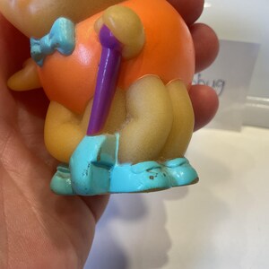 Puede incluir: Un peque&ntilde;o juguete de goma naranja y amarillo con un palo morado en la mano. El juguete lleva una pajarita azul y zapatos azules.