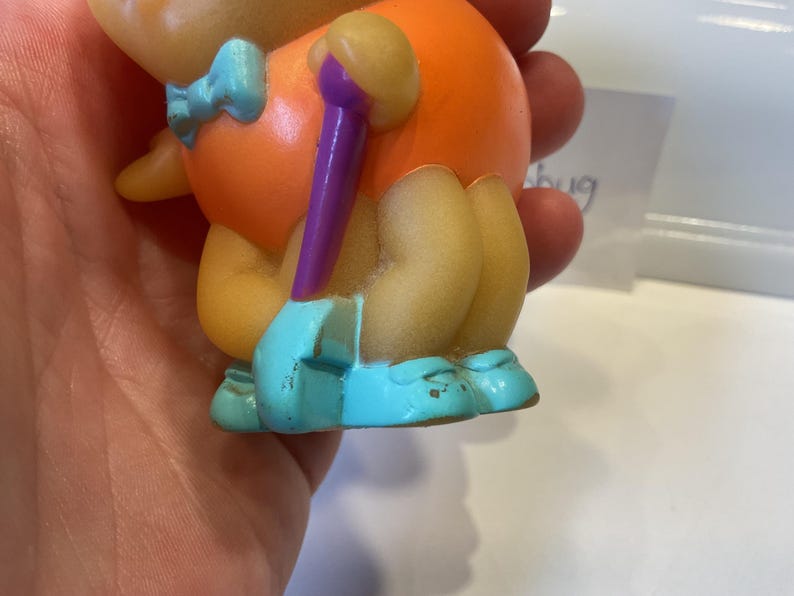 Puede incluir: Un peque&ntilde;o juguete de goma naranja y amarillo con un palo morado en la mano. El juguete lleva una pajarita azul y zapatos azules.