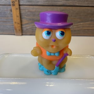 Puede incluir: Un juguete de goma amarillo con un sombrero morado, una camisa naranja y zapatos azules. El juguete sostiene un bast&oacute;n morado.