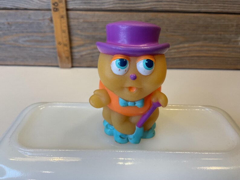 Puede incluir: Un juguete de goma amarillo con un sombrero morado, una camisa naranja y zapatos azules. El juguete sostiene un bast&oacute;n morado.