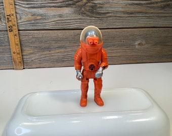 Könnte beinhalten: Orangefarbenes Astronautenspielzeug aus Plastik mit einem durchsichtigen Plastikhelm und silbernen Akzenten. Das Spielzeug steht auf einer weißen Oberfläche.
