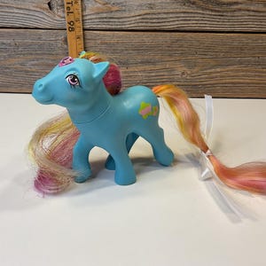 Op de afbeelding: Een vintage blauw speelgoedpony met een roze en gele manen en staart. De pony heeft een bloemdesign op zijn zij en een roze bloem op zijn hoofd. De pony is een verzamelobject.