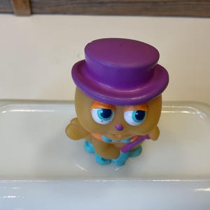 Puede incluir: Un peque&ntilde;o juguete de pl&aacute;stico amarillo con un sombrero morado y detalles azules y naranjas. El juguete tiene ojos azules grandes y una peque&ntilde;a nariz naranja.