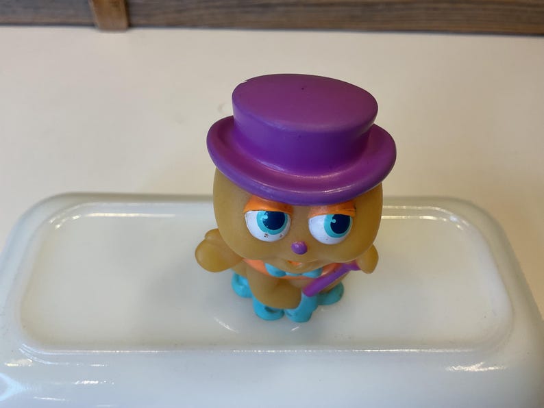 Puede incluir: Un peque&ntilde;o juguete de pl&aacute;stico amarillo con un sombrero morado y detalles azules y naranjas. El juguete tiene ojos azules grandes y una peque&ntilde;a nariz naranja.