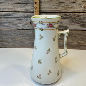 May include: A white ceramic pitcher with a floral design. The pitcher features a handle and a decorative band with pink roses and blue ribbons. Small floral sprigs are scattered across the body of the pitcher. The rim and handle are trimmed with green.