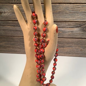 Puede incluir: Un collar largo hecho de cuentas de vidrio naranja con detalles azules y dorados. Las cuentas son redondas y tienen un patrón floral.
