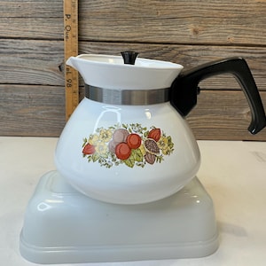 May include: A white teapot with a black handle and lid knob, featuring a central botanical illustration of red, orange, and brown fruits with green leaves. A metal band encircles the neck, and a wooden ruler is visible in the background.