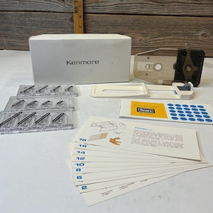 Puede incluir: Un kit de accesorios para máquina de coser Kenmore que incluye una caja blanca, un accesorio de plástico transparente, tres bandejas transparentes con piezas metálicas, una guía de plástico blanca, una tarjeta de instrucciones y tarjetas.
