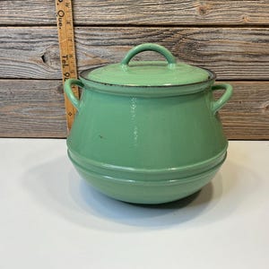 Puede incluir: Una olla vintage de esmalte verde claro con tapa y dos asas laterales. La olla tiene forma redondeada con un borde negro alrededor de la tapa y el borde. Una regla de madera es visible en el fondo.