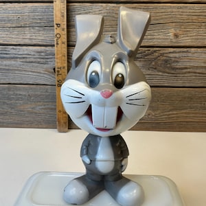 Puede incluir: Una figura vintage de Bugs Bunny. El personaje de dibujos animados está representado con un traje gris con una cara blanca, orejas grandes y una nariz rosa. La figura está sobre una base blanca, con una regla de madera en el fondo.