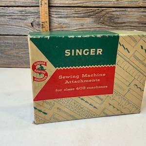 Puede incluir: Una caja de cartón rectangular para accesorios de máquina de coser Singer para máquinas de clase 403. La caja presenta un esquema de colores verde, rojo y beige con el logotipo de Singer y varias ilustraciones de patrones de costura impresas.