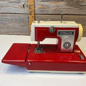 Puede incluir: Una máquina de coser Sewmate vintage roja y blanca con una mesa abatible. La máquina tiene un devanador de bobina blanco y un pedal de pie plateado.
