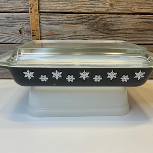 May include: A rectangular black Pyrex baking dish with a clear glass lid. The dish is decorated with white snowflake designs along the sides. It is sitting on a white rectangular base.