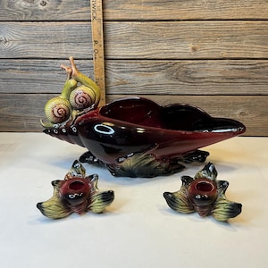 May include: A ceramic snail-shaped centerpiece with a deep burgundy interior and green and yellow snail details. Two matching leaf-shaped candle holders with red centers and green and yellow accents are also present.