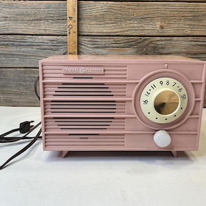 Könnte beinhalten: Ein altrosa General Electric Radio mit horizontalem Lamellen-Lautsprechergitter, einem großen, cremefarbenen, runden Abstimmknopf mit schwarzen Zahlen und einem kleinen weißen Lautstärkeregler. Ein braunes Stromkabel mit Stecker ragt links heraus.