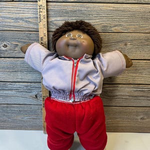 May include: A vintage Cabbage Patch Kid doll wearing a light purple zip-up jacket with a red zipper, red pants, and white shoes. The doll has brown hair and a smiling face. A wooden ruler is next to the doll.