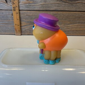Puede incluir: Un juguete de goma amarillo con un sombrero morado, una camisa naranja y zapatos azules. El juguete sostiene un palo morado.