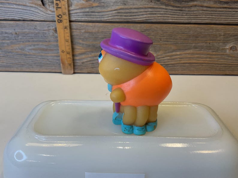 Puede incluir: Un juguete de goma amarillo con un sombrero morado, una camisa naranja y zapatos azules. El juguete sostiene un palo morado.