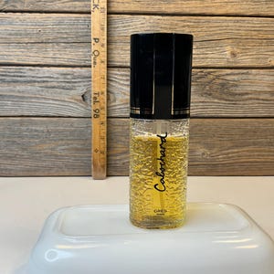 May include: A cylindrical glass perfume bottle with a textured surface containing a golden liquid. The bottle features a black cap and the text "Cabochard" and "GRES PARIS" printed on the front. A wooden ruler is visible in the background.
