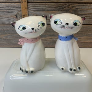 Könnte beinhalten: Zwei weiße Keramik-Katzenfiguren mit bemalten blauen und rosa Schleifenakzenten. Die Katzen sitzen auf einer weißen Keramikbasis.