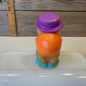 Puede incluir: Una peque&ntilde;a figura de juguete de goma naranja y amarilla que lleva un sombrero morado y zapatos azules. La figura est&aacute; de pie sobre una superficie blanca.