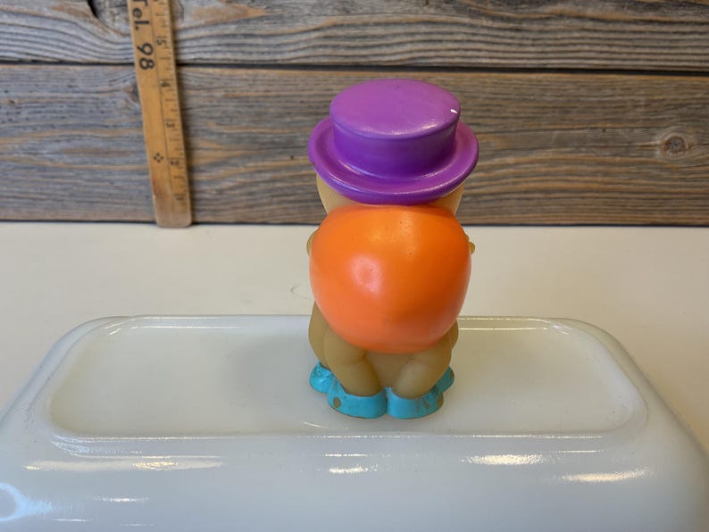 Puede incluir: Una peque&ntilde;a figura de juguete de goma naranja y amarilla que lleva un sombrero morado y zapatos azules. La figura est&aacute; de pie sobre una superficie blanca.