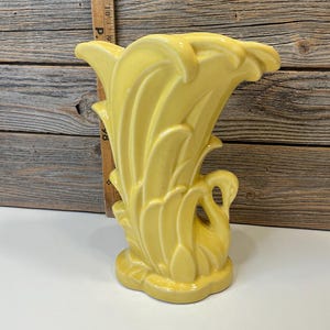 Op de afbeelding: Een vintage gele keramische vaas met een zwanenontwerp. De vaas heeft een uitlopende bovenkant met bladvormige details en een zwanenfiguur aan de zijkant. De vaas is lichtgeel van kleur en heeft een glanzende afwerking. De basis heeft een afgeronde vorm.