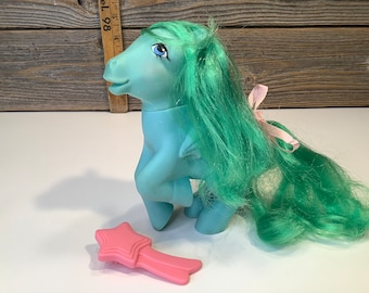 May include: A blue and green My Little Pony toy with a pink bow and a pink star-shaped brush. The pony has long green hair and blue eyes.
