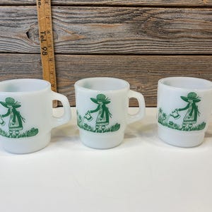 Puede incluir: Tres tazas de cerámica blanca con una ilustración verde de una persona regando un jardín. Las tazas tienen un asa y una base ligeramente acampanada. La ilustración se repite en cada taza. Altura aproximada: 8 cm.