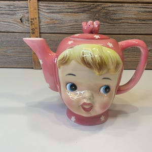May include: A pink ceramic teapot shaped like a girl's head with white flowers and a pink bird on the lid.